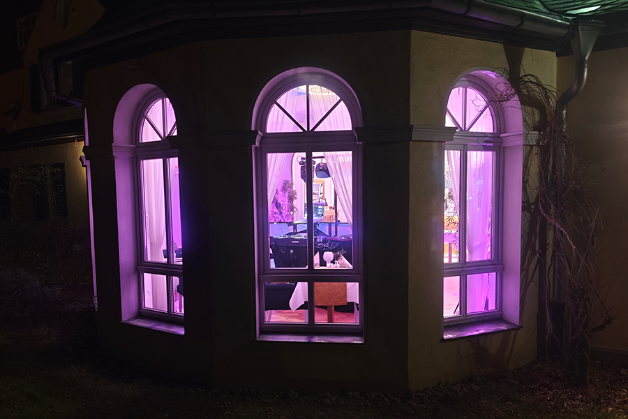 Abendlicher Blick von außen durch große Rundbogenfenster in eine stimmungsvoll beleuchtete Eventlocation mit violetter Ambientebeleuchtung und festlich gedecktem Tisch.