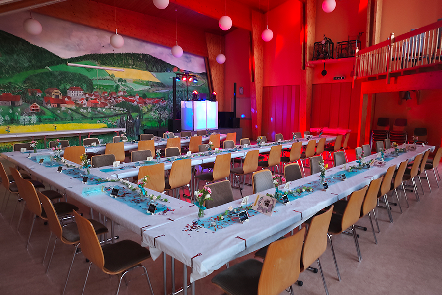 estlich dekorierter Veranstaltungsraum mit U-förmig gedeckten Tischen, liebevoller Tischdekoration, moderner Lichttechnik und DJ-Setup vor einer großflächigen Wandmalerei mit Dorfmotiv.
