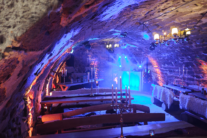 Stimmungsvolle Eventlocation in einem urigen Gewölbekeller mit atmosphärischer Beleuchtung, festlich gedeckten Holzbänken, Kerzenleuchtern und professioneller Lichttechnik im Hintergrund