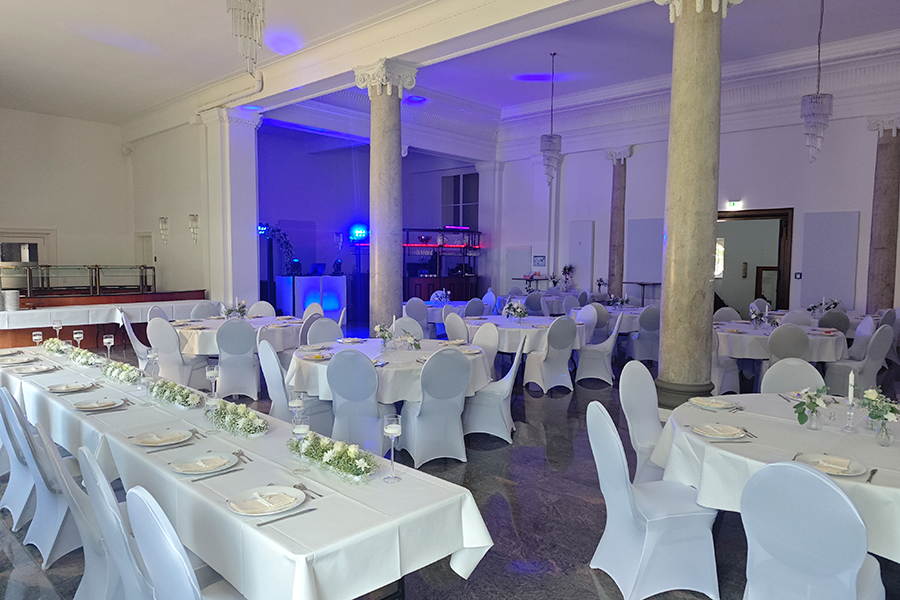 Weiß gedeckter Hochzeitssaal mit runden Tischen, Blumenschmuck und DJ-Technik im Hintergrund bei einer Hochzeit mit Hochzeits-DJ Andreas.