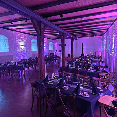 Stimmungsvoll beleuchteter Hochzeitssaal mit rustikalen Steinwänden, Holzbalkendecke, langen gedeckten Tafeln und violett-blauer Ambientebeleuchtung sowie DJ-Setup im Hintergrund.