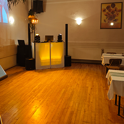 Kleiner Festsaal mit hellem Holzboden, gelb beleuchtetem DJ-Pult, Lautsprechern, Moving-Head-Lichtern und eingedeckten Tischen am Rand.