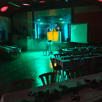 Festsaal mit eingedeckten Tischen, grün-blauer Partybeleuchtung, Nebeleffekten und beleuchteter DJ-Bühne mit Leinwand, vorbereitet für eine Geburtstagsfeier.