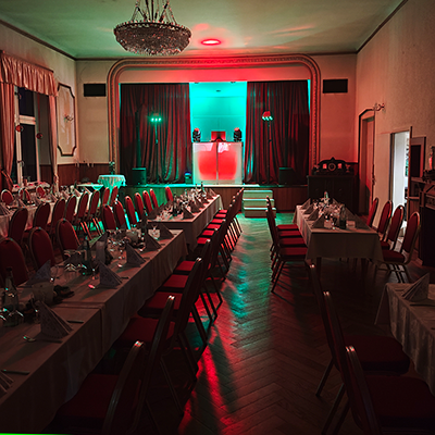 Großer Festsaal mit eingedeckten Tischen, roten Stühlen und beleuchteter DJ-Bühne in Rot und Grün, vorbereitet für eine Firmenweihnachtsfeier.