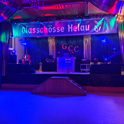 Farbig ausgeleuchtete Karnevalsbühne mit DJ-Pult, Lichttechnik, Instrumenten und großem „Glasschösse Helau“-Banner in dekorierter Veranstaltungshalle.