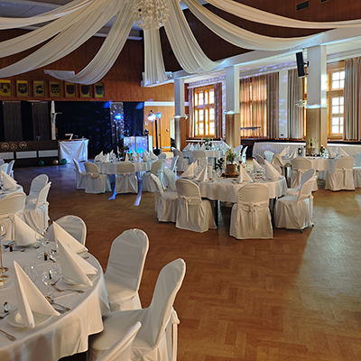 Ein großer Hochzeitssaal in Mühlhausen mit Parkettboden und runden, weiß gedeckten Tischen. Weiße Stoffbahnen drapieren die hohe Decke. Im Hintergrund ist das DJ-Setup von DJ Andreas mit weißem Pult und Lichttechnik vor einer dunklen Trennwand mit Wappen-Dekoration aufgebaut.