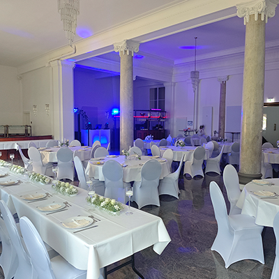 Ein großer Festsaal in Mühlhausen, festlich dekoriert für eine Hochzeit. Weiße Stuhlhussen mit türkisfarbenen Schleifen ergänzen die blau-türkise Ambientebeleuchtung an den Wänden. Im Hintergrund ist ein weißes DJ-Pult mit Säulenlautsprechern und Moving-Heads vor einer großen Fensterfront positioniert.