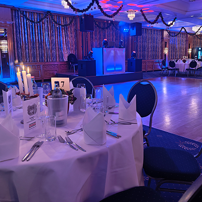 Ein Festsaal mit blauem Teppich, gedeckten runden Tischen, Tannengirlanden und einer blau beleuchteten Bühne mit DJ-Pult für eine Firmenweihnachtsfeier in einem Hotel mit Event-DJ Buttstädt.