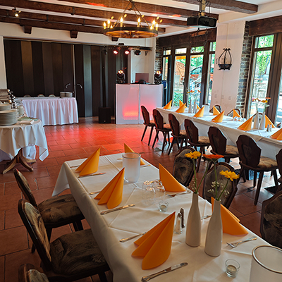 Ein Raum mit Terrakotta-Fliesen, Holzbalken, gedeckten Tischen mit orangen Servietten und rot beleuchtetem DJ-Pult, bereit für eine Silberhochzeit in einem Landgasthof mit Event-DJ aus Sömmerda.