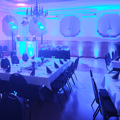 Ein blau illuminierter Saal mit Holzvertäfelung, gedeckten Tischen mit schwarzen Servietten, einer Buffetstation und einem DJ-Pult, bereit für eine Silvesterfeier mit DJ Sömmerda.