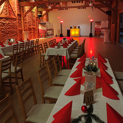 Ein rustikaler Saal mit Backsteinwänden, gedeckten Tischen mit roten Servietten, Tannengrün und einem beleuchteten DJ-Pult, bereit für eine Weihnachtsfeier mit Event-DJ Buttstädt.