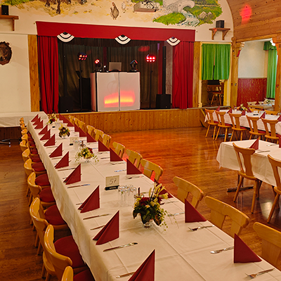 Ein rustikaler Saal mit Holzboden, langen gedeckten Tafeln, roten Servietten und einer Bühne mit beleuchtetem DJ-Pult für eine Firmenfeier mit DJ Bad Langensalza in einem Landhotel.