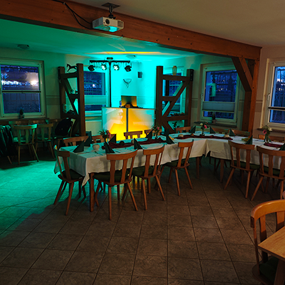 Ein Veranstaltungsraum mit Fliesenboden, gedeckten Tischen, Holzstühlen und einem gelb-grün beleuchteten DJ-Pult, vorbereitet für das Event: kleiner 65. Geburtstag mit DJ Bad Langensalza in einer rustikalen L